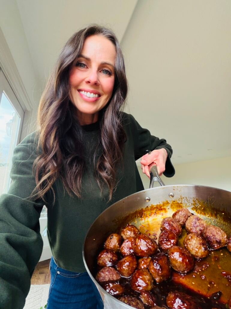 Carrian holding a large skillet with browned meatballs coated in a sticky sauce
