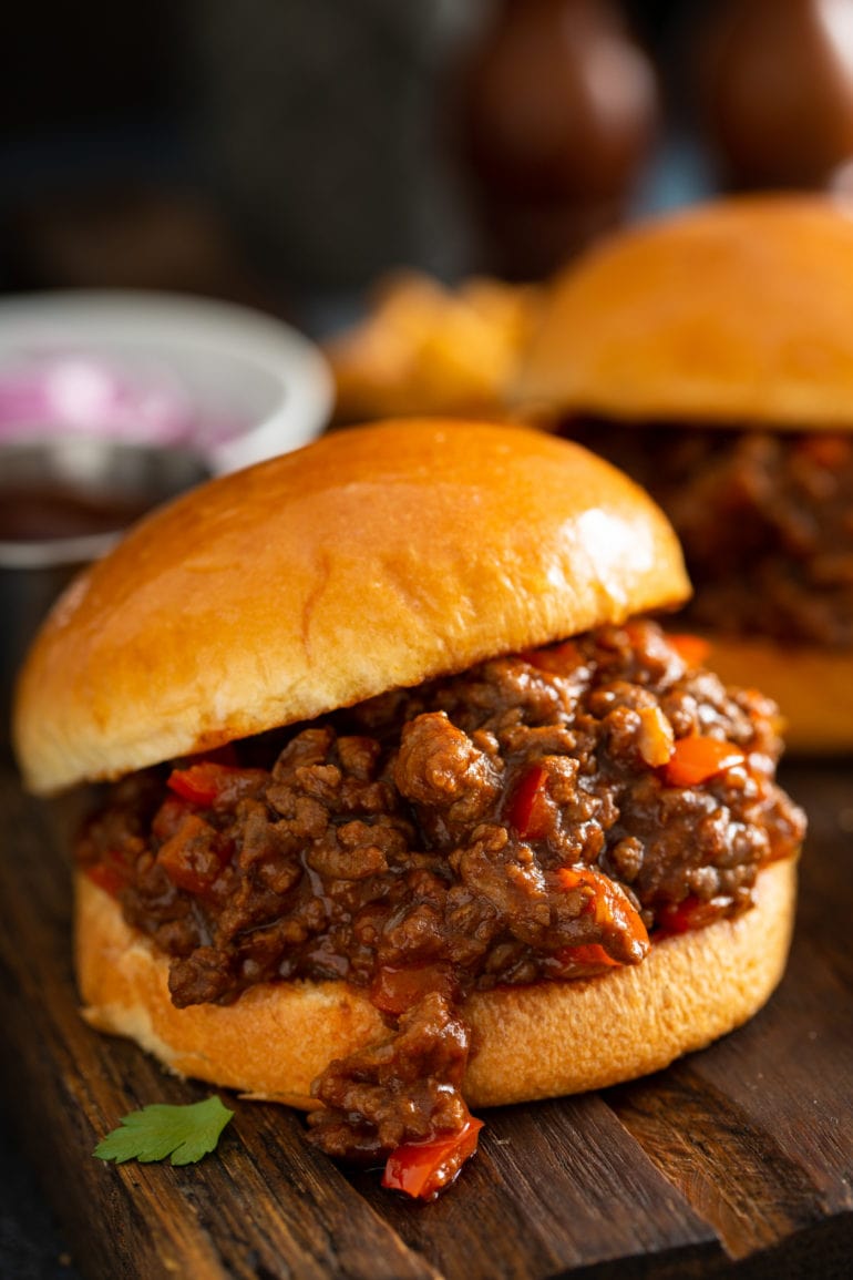 Sloppy joe on a toasted bun. The sloppy joe has browned hamburger and chopped red bell peppers and is thick and saucy. Another sloppy joe and a small bowl of sliced onions is in the background.