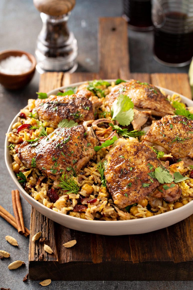 A small dish of salt and a crystal pepper grinder are the background to a large bowl of Jerusalem rice topped with Jerusalem chicken thighs that have been sprinkled with cilantro and dill leaves. There are cinnamon sticks on the table next to the bowl.