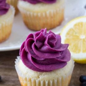 Moist lemon cupcake topped with blueberry frosting with a a plate of lemon blueberry cupcakes in the background.