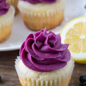 Moist lemon cupcake topped with blueberry frosting with a a plate of lemon blueberry cupcakes in the background.