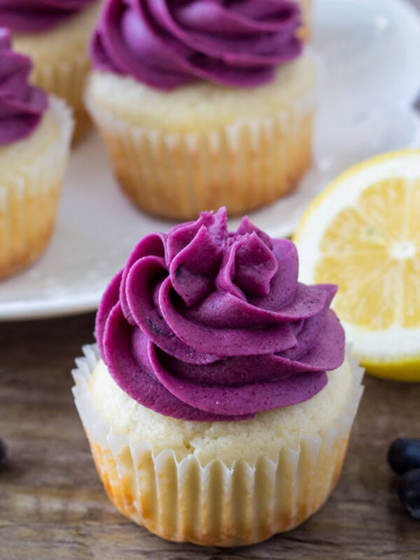 Moist lemon cupcake topped with blueberry frosting with a a plate of lemon blueberry cupcakes in the background.