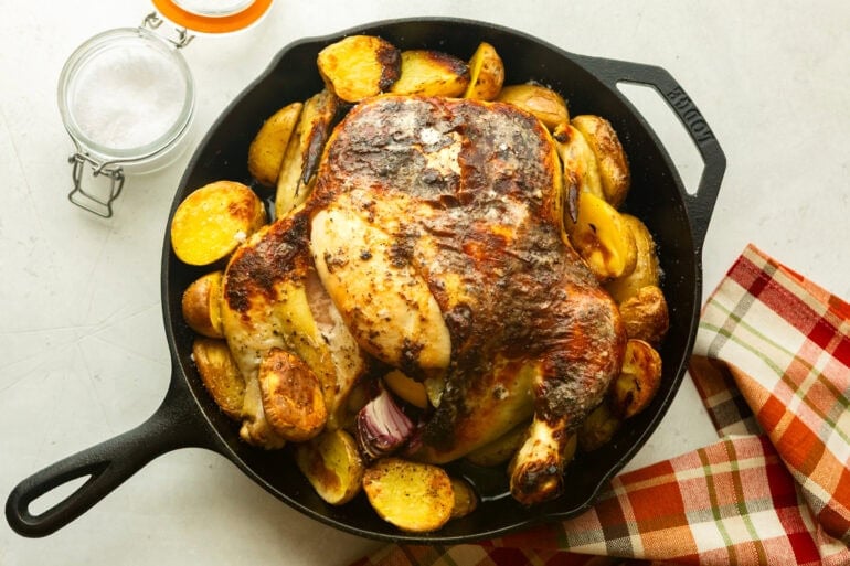 a large cast iron skillet full of a whole roasted chicken surrounded by golden crispy potatoes