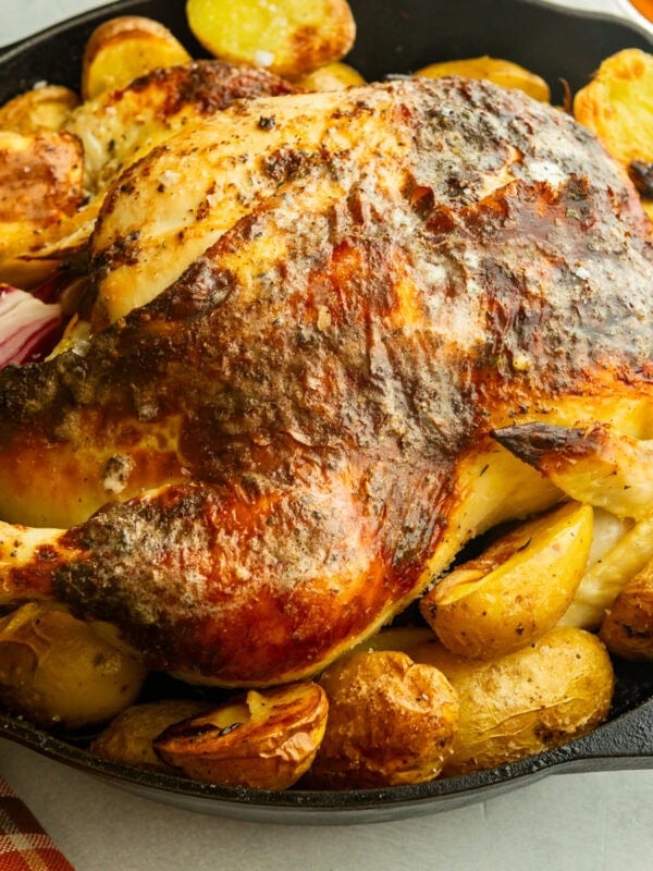 a large cast iron skillet with a roasted whole chicken in it surrounded by golden roasted potatoes