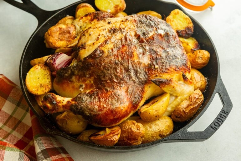 a large cast iron skillet with a roasted whole chicken in it surrounded by golden roasted potatoes