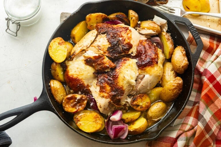 a whole roasted chicken with golden skin peeling off it surrounded by roasted potatoes all in a large cast iron skillet