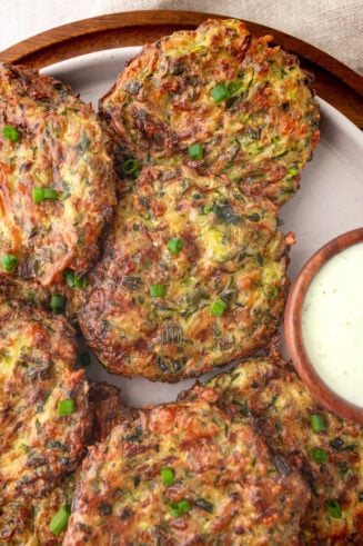 a photo of several golden fried zucchini fritters sitting next to a small cup of creamy dipping sauce