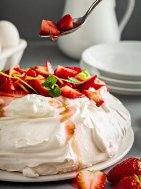 Pavlova on a serving plate. It is topped with whipped cream, sliced strawberries, slivers of lemon rind, and a few mint leaves. A spoon with strawberries and juice drizzling down is above the pavlova. There are white dessert plates, eggs, and a white pitcher are in the background.