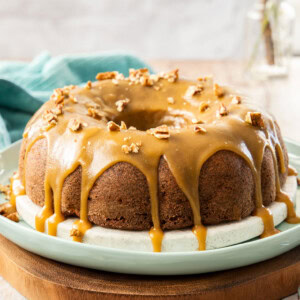 A bundt cake with caramel icing dripping down it, on a white and green platter. It sits on a wooden board