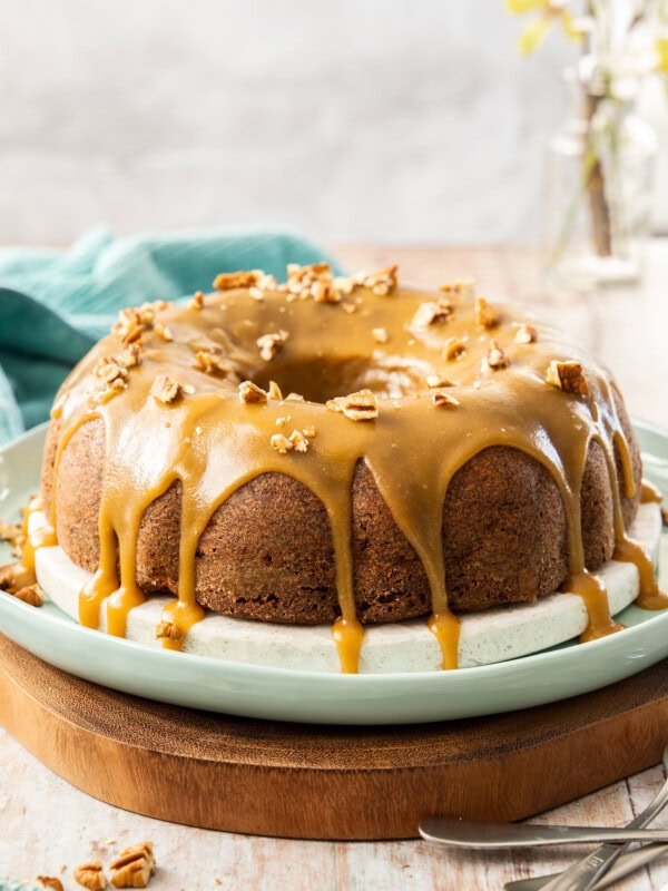 A bundt cake with caramel icing dripping down it, on a white and green platter. It sits on a wooden board