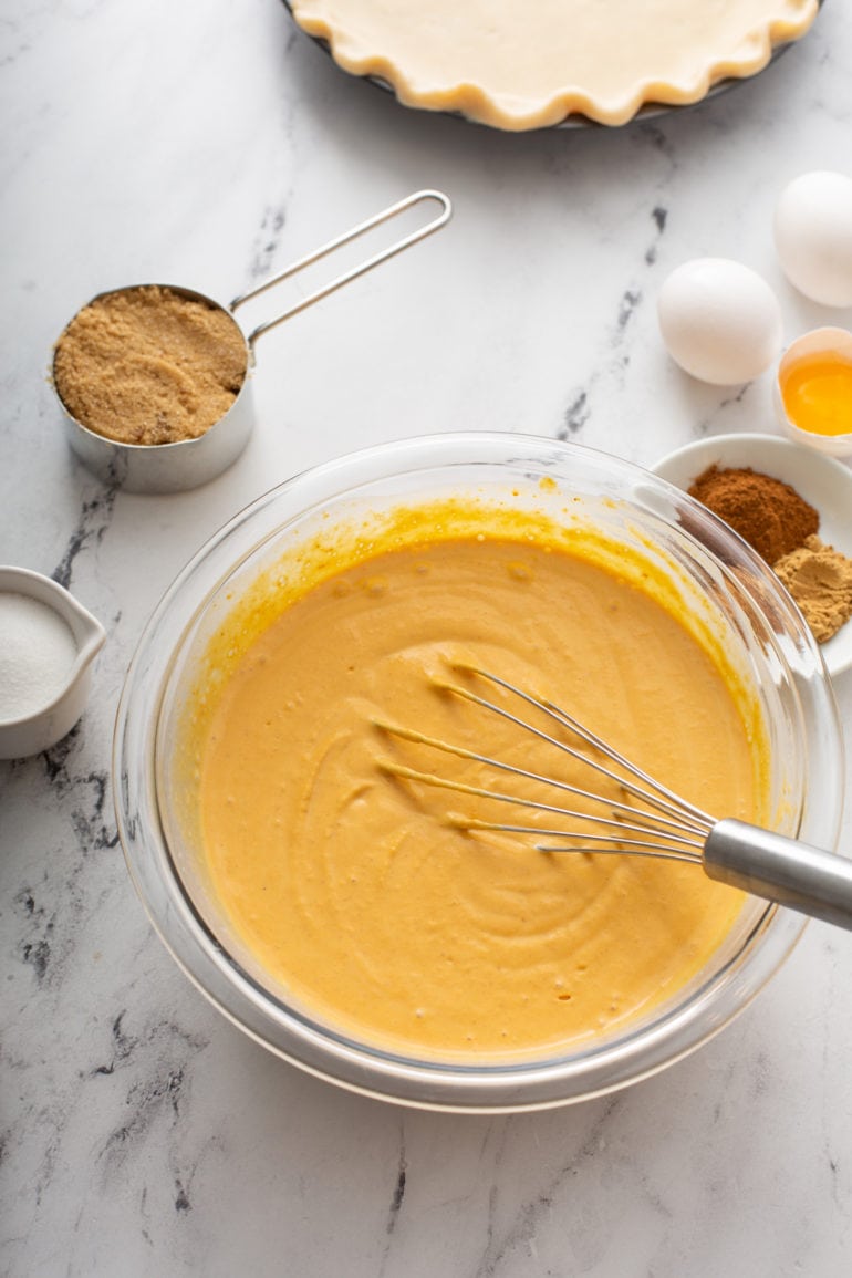 A photo of a glass bowl full of pumpkin pie filling surrounded by ingredients for pumpkin pie.