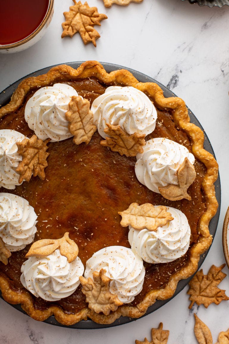 A photo of a pumpkin pie topped with piped whipped cream and stamped pie crust leaves taken from above.