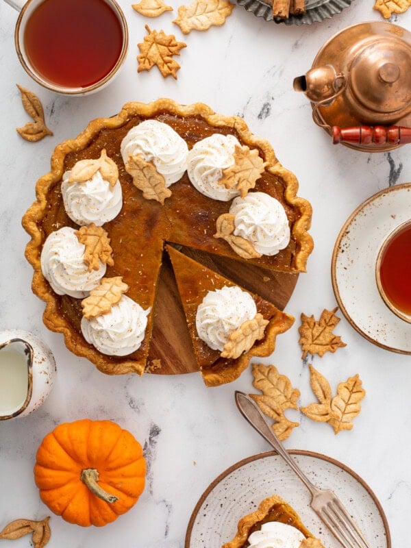 A photo of a pumpkin pie topped with piped whipped cream and stamped pie crust leaves taken from above with a slice pulled out.
