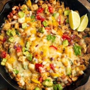 a cast iron skillet full of golden chicken chunks, melted cheese, avocado, zucchini and a couple of lime wedges