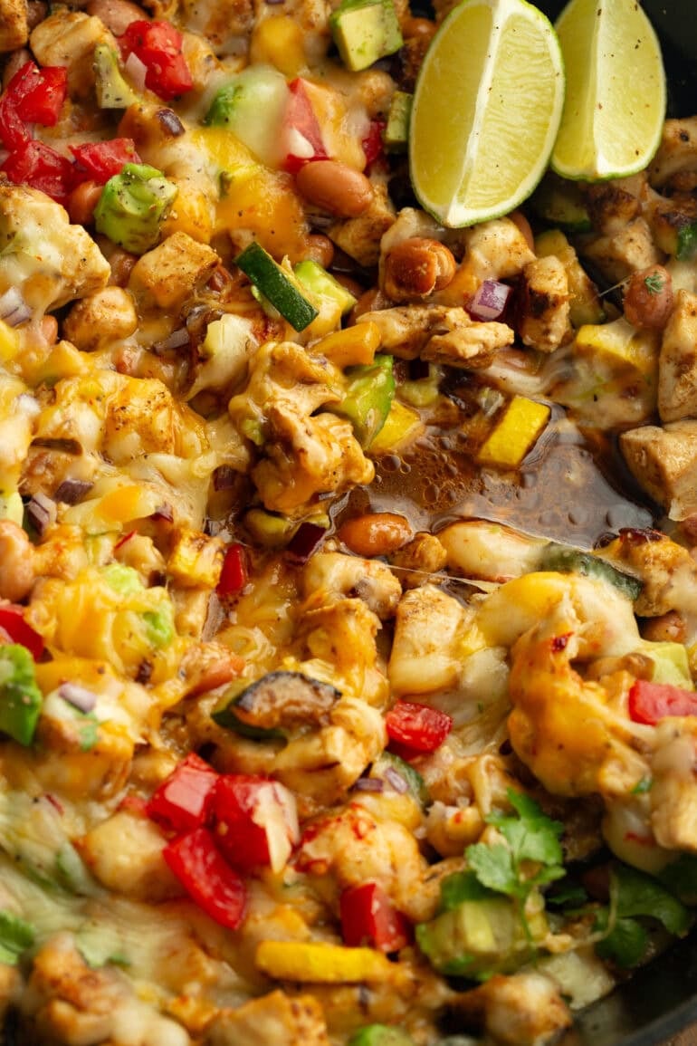 close up of golden chunks of chicken sauteed with zucchini and yellow squash and topped with melted cheese, avocado, and tomatoes
