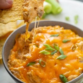 A bowl of buffalo chicken dip. A chip has been dipped in and melty stringy cheese is hanging from the tip of the chip. Chips and celery sticks are in the background.