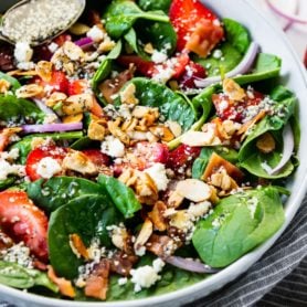 a photo of a large white serving bowl full of a spinach salad topped with sliced strawberries, sliced red onions, candied slivered almonds, feta cheese, bacon crumbles and homeade poppy seed dressing.