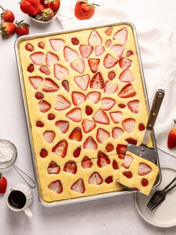 a half sheet pan full of baked sheet pan pancakes topped with slices of fresh strawberry baked into the pancakes and a metal spatula removing one serving