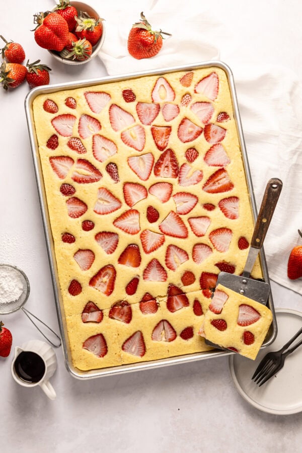 a half sheet pan full of baked sheet pan pancakes topped with slices of fresh strawberry baked into the pancakes and a metal spatula removing one serving