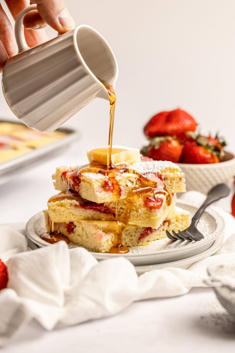 someone pouring maple syrup over a stack of strawberry sheet pan pancakes 