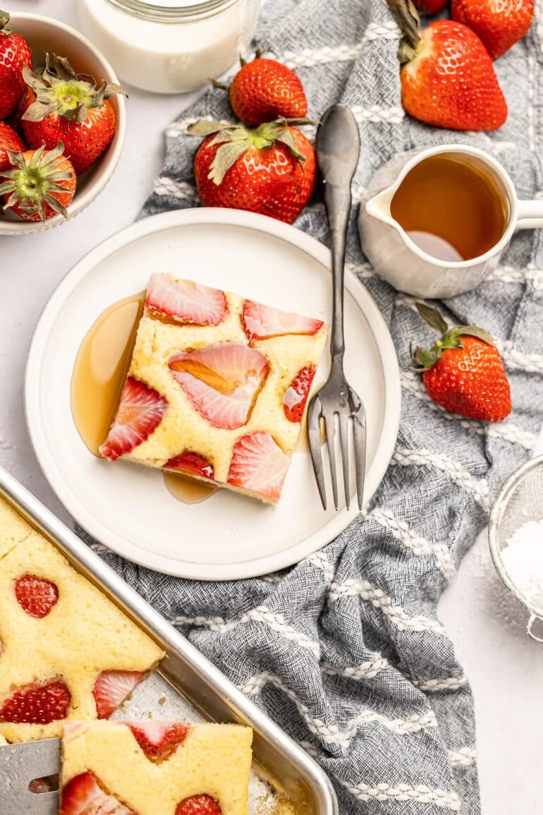 a serving of sheet pan pancakes on a small plate topped with syrup with a bottle of syrup on the side and a sheet pan of sheet pan pancakes sitting next to it