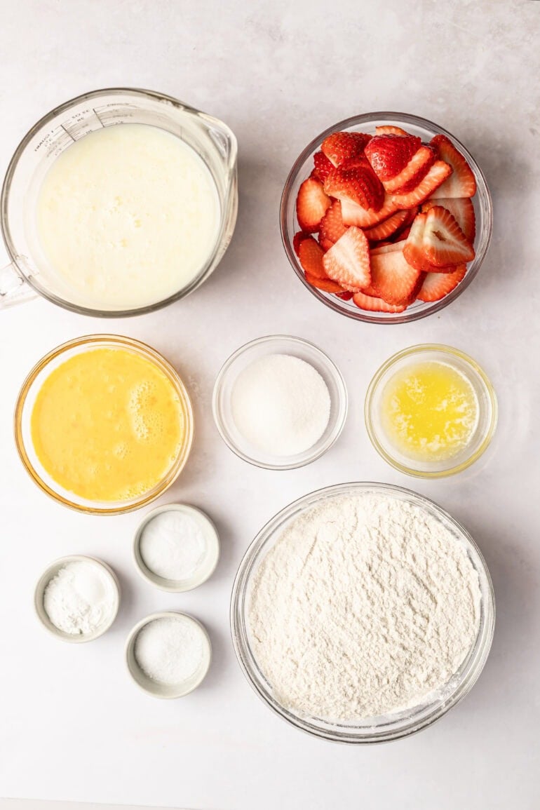 all the ingredients for sheet pan pancakes in separate small glass bowls including flour, buttermilk, sugar, eggs, baking powder and soda, melted butter and sliced strawberries