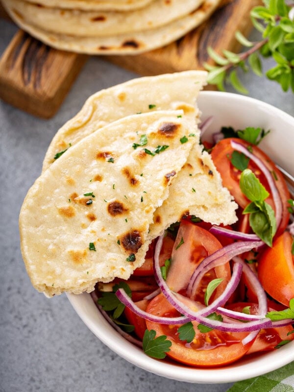 A photo of a bowl of fresh tomato and onion salad in a white bowl with torn pieces of unleavened bread sitting on the side of the bowl.