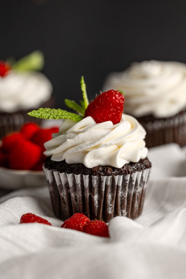a photo of a chocolate cupcake in a cupcake liner topped with piped cream cheese frosting with a single raspberry and couple mint leaves on top
