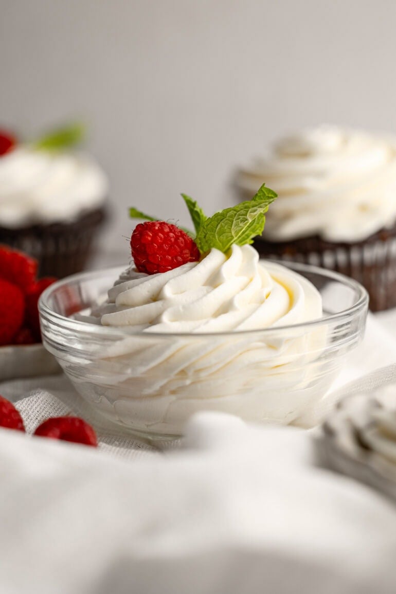 a photo of a small glass bowl full of piped whipped cream cream cheese frosting with a single raspberry on top and a few mint leaves