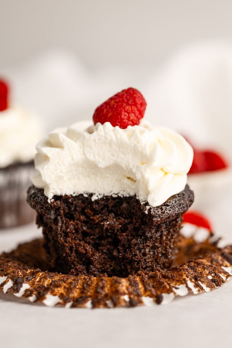 a photo of a chocolate cupcake topped with thick cream cheese frosting with a bite taken out of it and a single raspberry sitting on top