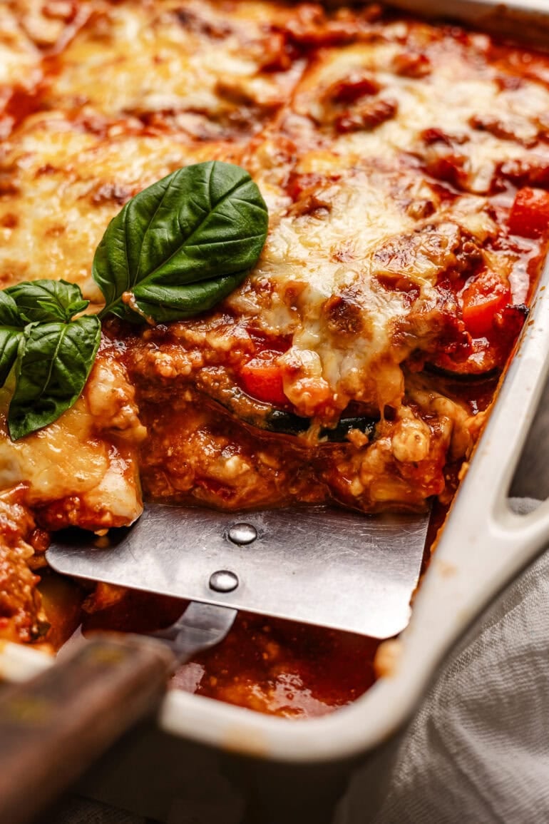 a metal spatula is digging into a full baking dish of layered veggies, meat sauce, and cheese.