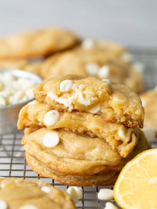 a photo of a stack of golden lemon cheesecake cookies full of white chocolate chips with the top cookie broken in half with the insides of cookie showing.