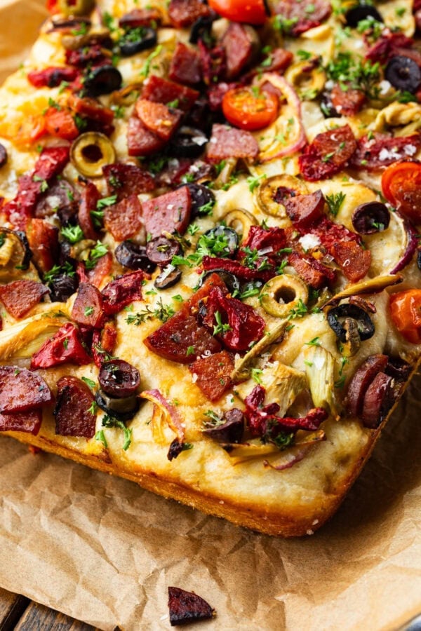 a baked loaf of antipasto focaccia bread topped with sliced green olives, chopped meats, sundried tomatoes and herbs