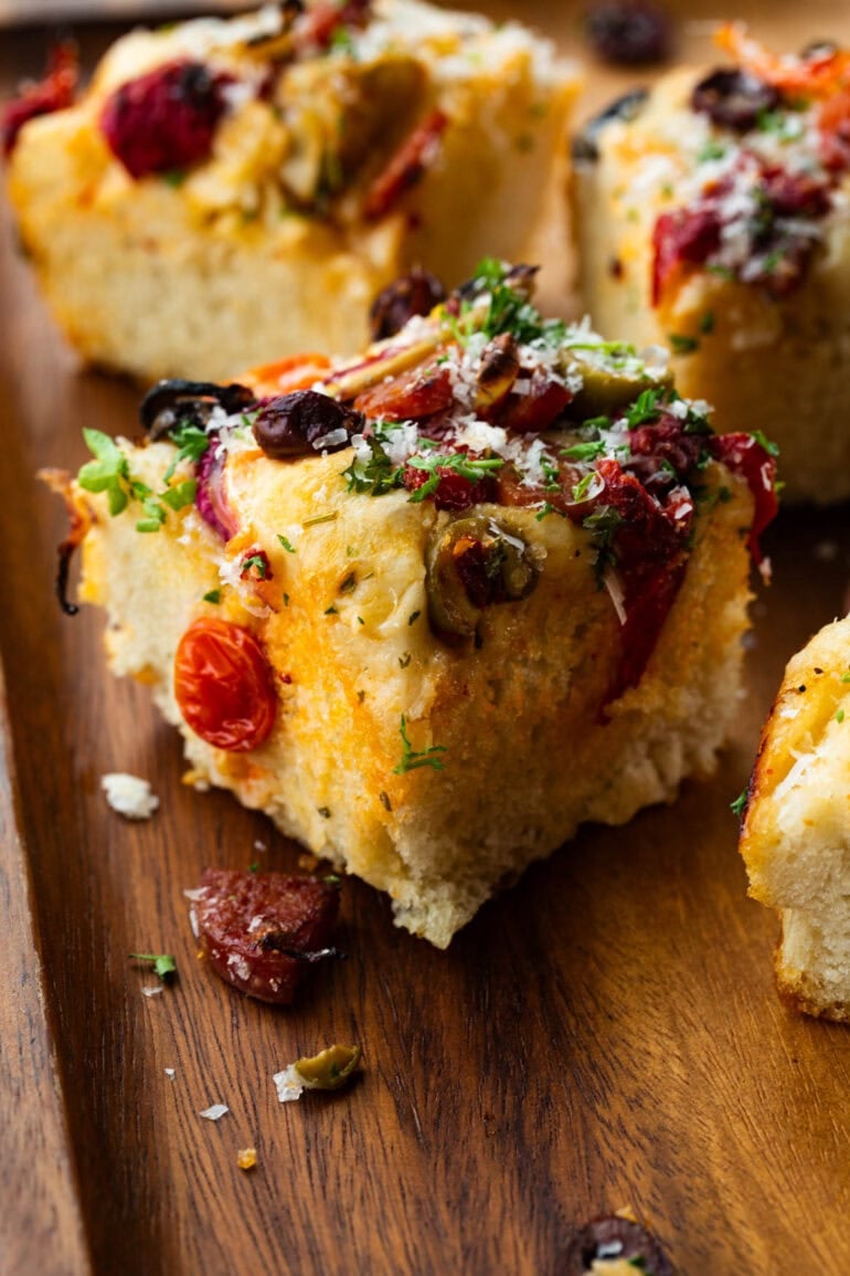 a piece of fluffy focaccia bread topped with antipasto including tomatoes, olives, chopped meats and artichoke hearts