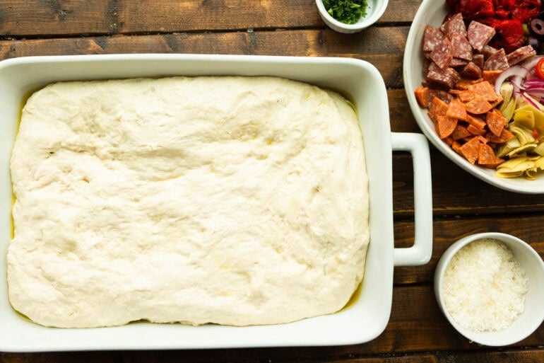 a casserole dish with unbaked focaccia bread dough in to with bowls of antipasto toppings around it