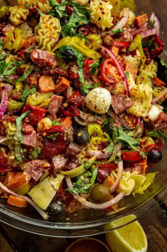 a photo of an antipasto pasta salad in a large glass bowl including cooked pasta, mozzarella pearls, pepperoncinis, salami, pepperoni, and olives