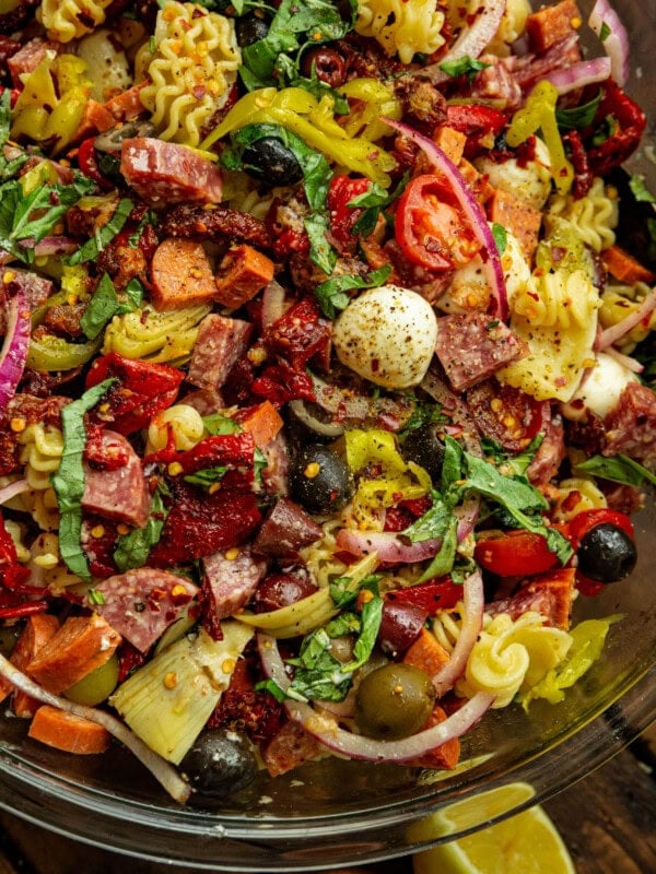 a photo of an antipasto pasta salad in a large glass bowl including cooked pasta, mozzarella pearls, pepperoncinis, salami, pepperoni, and olives