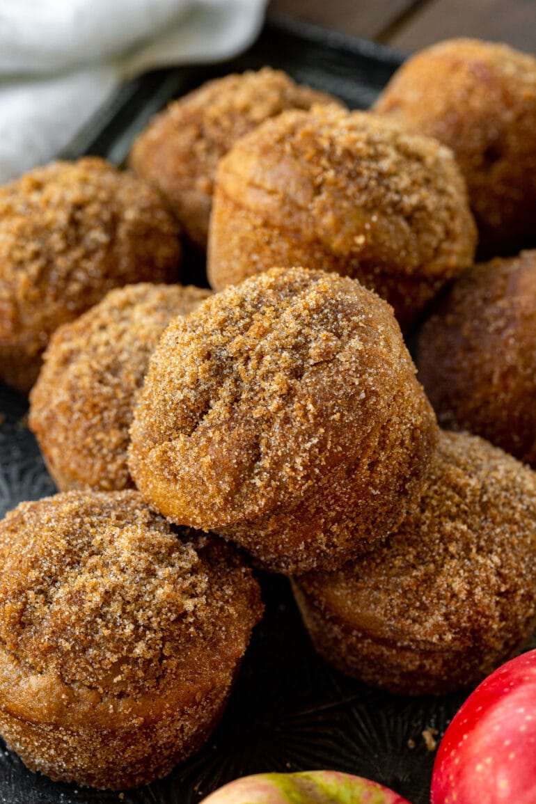 a photo of several golden baked apple cider donut muffins piled on each other and they are all topped with a crunchy brown sugar cinnamon topping.