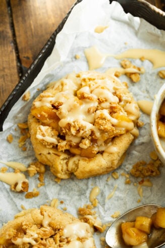 a photo of an apple crisp cookie with a golden cookie base topped with cookie apples, a crunchy oat topping and a vanilla glaze drizzle.
