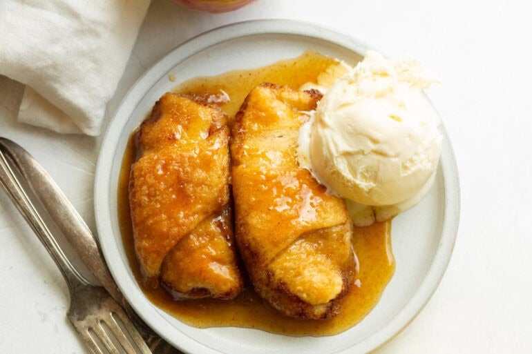 Golden crescent roll apple dumplings filled with cinnamon apples and baked in brown sugar sauce served with a scoop of vanilla ice cream.