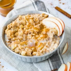 A bowl of slow cooker apple pie oatmeal. Caramel is drizzled over the top of the oatmeal and three slices of apple are on the edge of the oatmeal. More slices of apples and a cinnamon stick and a container of caramel are in the background.