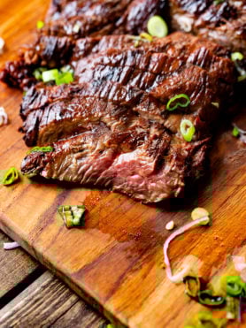 a photo of tender and juicy slices of flank steak sitting on a wooden cutting board topped with chopped scallions.