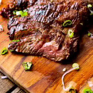 a photo of tender and juicy slices of flank steak sitting on a wooden cutting board topped with chopped scallions.
