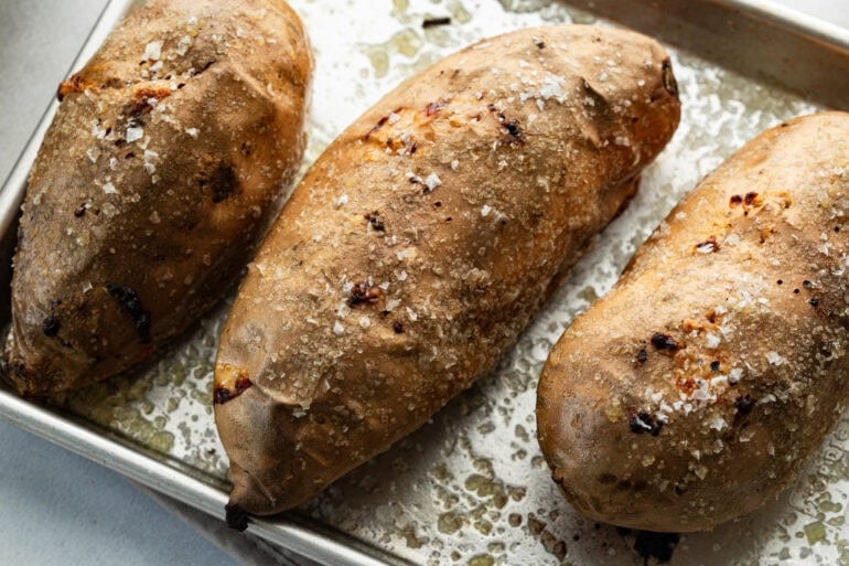 several whole white sweet potatoes on a baking sheet and sprinkled with sea salt flakes