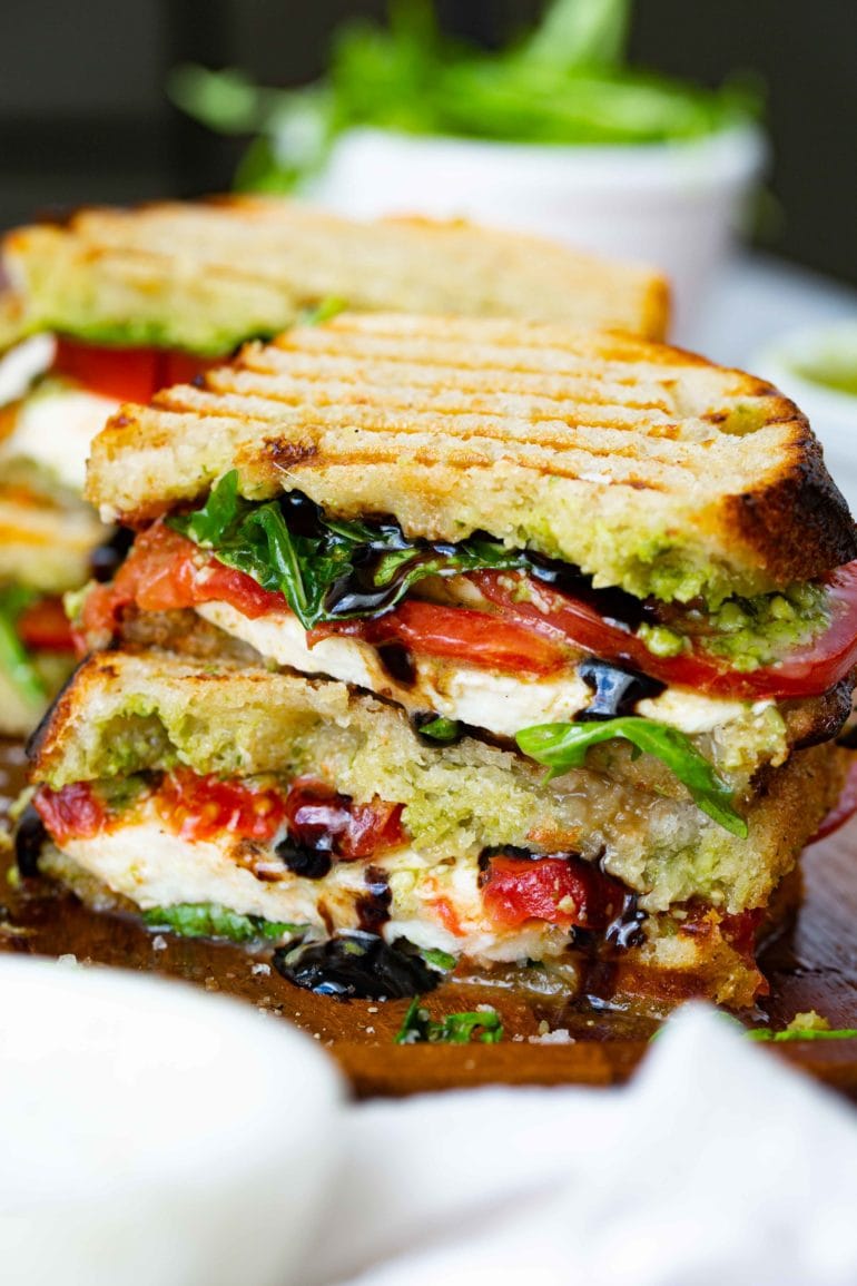 A balsamic glazed caprese sandwich that has been cut in half and stacked on top of each other. The sandwich has grill marks on top and has arugula, fresh mozzarella cheese slices and tomato slices inside. You can see the balsamic glaze dribbling out of the sandwich and another sandwich in the background.