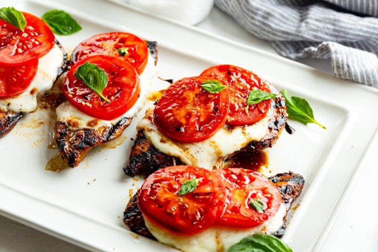 Several Capri chicken breasts on a plate topped with melted mozzarella, tomato slices, fresh basil and a drizzle of balsamic vinegar.