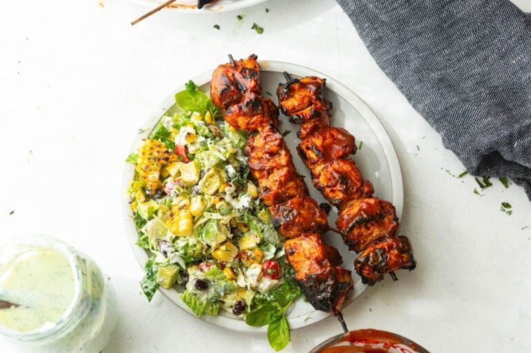 a white dinner plate with two grilled bbq chicken skewers with creamy ranch salad on the side