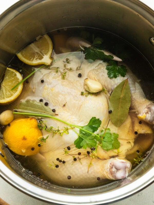 a large stock pot with an uncooked whole chicken in it submerged in brining liquid with halved lemons and herbs and peppercorns floating on top