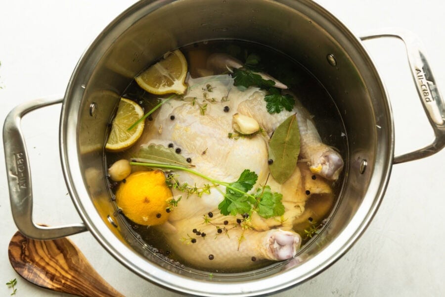 a large stock pot with an uncooked whole chicken in it submerged in brining liquid with halved lemons and herbs and peppercorns floating on top