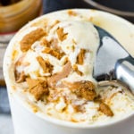 A photo of several scoops of Biscoff cookie butter homemade ice cream in a dark gray bowl with cookie crumbles and melted cookie butter all over the top.